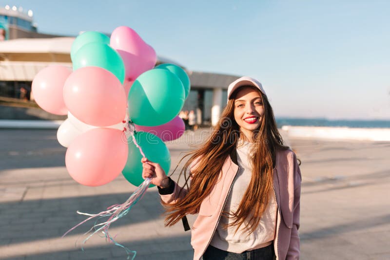 Portrait of beautiful long-haired brunette girl posing at the sea pier while wind waving her hair. Adorable young woman in trendy pink clothes standing with bunch of party balloons at the ocean wharf. Portrait of beautiful long-haired brunette girl posing at the sea pier while wind waving her hair. Adorable young woman in trendy pink clothes standing with bunch of party balloons at the ocean wharf