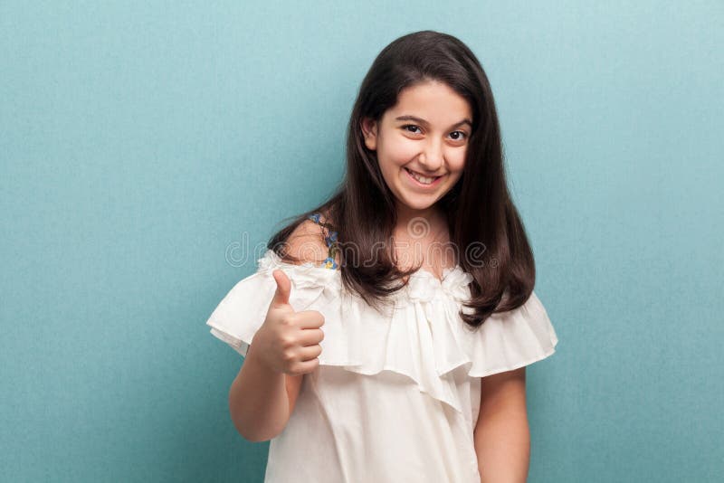Retrato da moça moreno bonita satisfeita feliz com cabelo reto longo preto na posição branca do vestido, polegares acima e foto de stock Retrato da moça moreno bonita satisfeita feliz com cabelo reto longo preto na posição branca do vestido, polegares acima e foto de stock