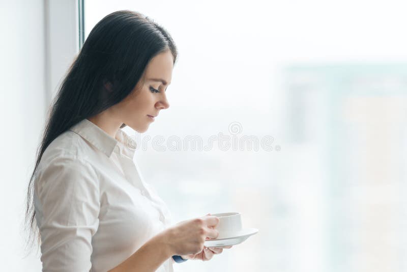 Retrato da mulher de negócios nova na camisa branca com xícara de café, mulher de sorriso que aprecia seu café aromático da manhã imagem de stock Retrato da mulher de negócios nova na camisa branca com xícara de café, mulher de sorriso que aprecia seu café aromático da manhã imagem de stock
