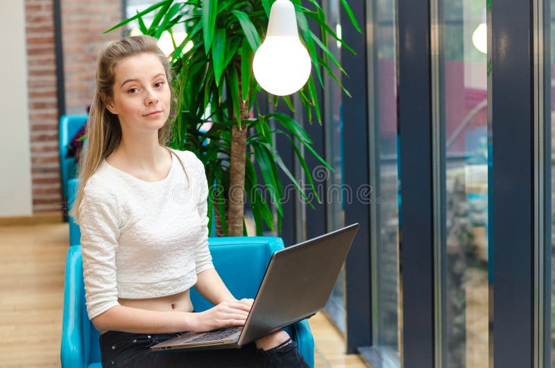 Retrato da mulher de sorriso bonita que senta-se em uma cadeira confortável em um café com portátil preto Estudante bonito que fa foto de stock royalty free Retrato da mulher de sorriso bonita que senta-se em uma cadeira confortável em um café com portátil preto Estudante bonito que fa foto de stock royalty free