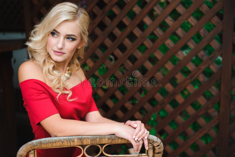 Retrato da mulher loura bronzeada bonita nova no assento vermelho do vestido de nivelamento exterior no café da rua apenas fotografia de stock Retrato da mulher loura bronzeada bonita nova no assento vermelho do vestido de nivelamento exterior no café da rua apenas fotografia de stock