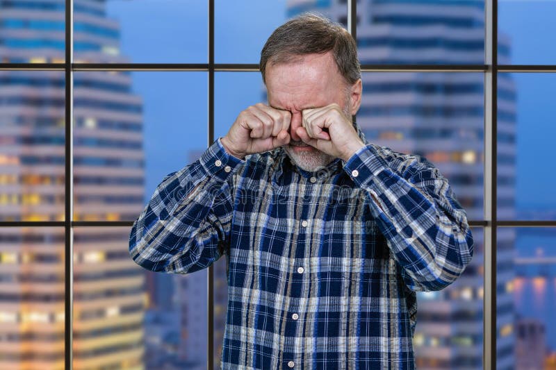 Portrait of crying aged senior man is wiping his tears. Checkered windows with night cityscape background. Portrait of crying aged senior man is wiping his tears. Checkered windows with night cityscape background.