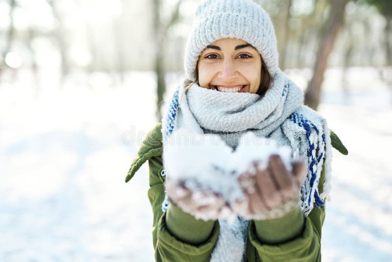Retrato de close de uma sorridente engraçada de chapéu de lã e longo cachecol quente segurando neve para a câmera no inverno de n fotografia de stock Retrato de close de uma sorridente engraçada de chapéu de lã e longo cachecol quente segurando neve para a câmera no inverno de n fotografia de stock