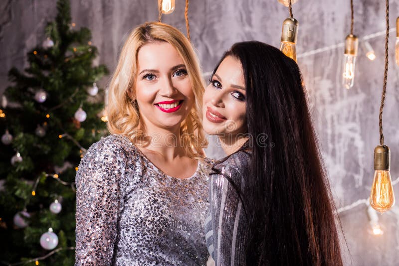 Retrato de dos mujeres bonitas preciosas en los vestidos brillantes que abrazan y que miran la cámara aislada sobre el árbol de n imagenes de archivo Retrato de dos mujeres bonitas preciosas en los vestidos brillantes que abrazan y que miran la cámara aislada sobre el árbol de n imagenes de archivo