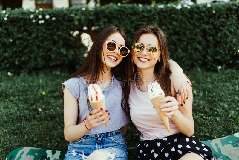Retrato de dos mujeres jovenes que se colocan junto de consumición del helado que abraza sentarse en la hierba en calle de la ciu fotografía de archivo libre de regalías Retrato de dos mujeres jovenes que se colocan junto de consumición del helado que abraza sentarse en la hierba en calle de la ciu fotografía de archivo libre de regalías