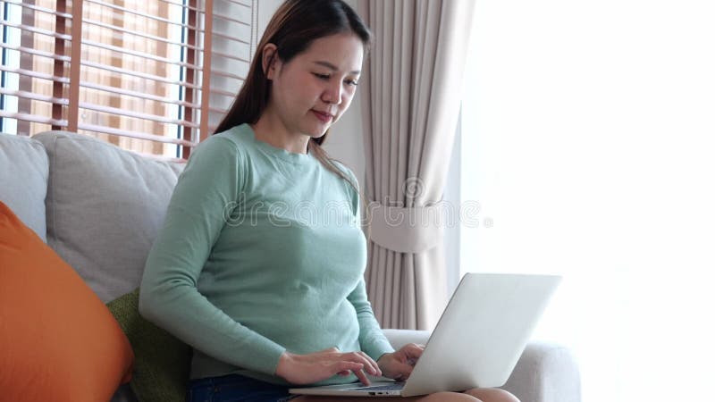 Retrato de joven mujer de negocios asiática sentada en un sofá usando computadora portátil en la sala de estar en casa. almacen de metraje de vídeo