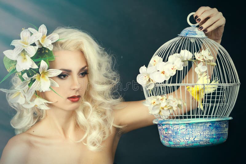 Beauty portrait of a girl with a lily in her hair. Blonde holding a cage with a bird on a gray background. Beauty portrait of a girl with a lily in her hair. Blonde holding a cage with a bird on a gray background