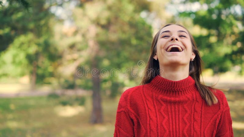 Retrato de la cámara lenta de la muchacha alegre en el suéter caliente que se coloca en el parque con sonrisa alegre, riendo y mi almacen de metraje de vídeo