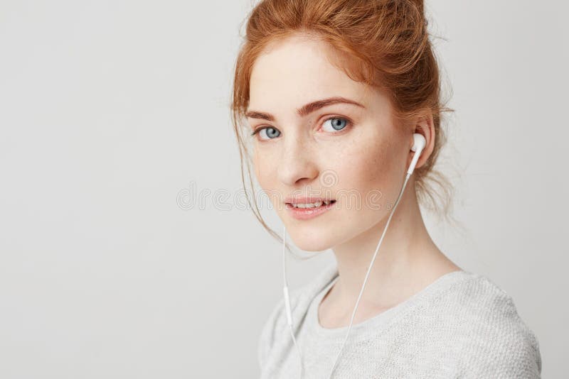 Retrato de la muchacha blanda hermosa joven del pelirrojo con los ojos azules en los auriculares que miran la cámara que sonríe s imagen de archivo libre de regalías Retrato de la muchacha blanda hermosa joven del pelirrojo con los ojos azules en los auriculares que miran la cámara que sonríe s imagen de archivo libre de regalías
