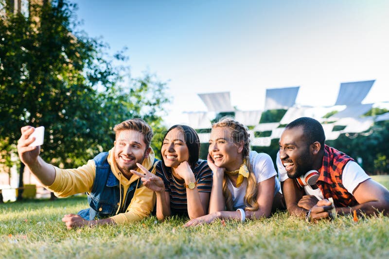 retrato de los amigos alegres multiétnicos que toman el selfie en smartphone mientras que descansa sobre hierba verde fotos de archivo libres de regalías retrato de los amigos alegres multiétnicos que toman el selfie en smartphone mientras que descansa sobre hierba verde fotos de archivo libres de regalías