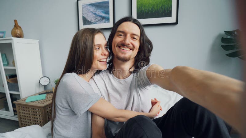 Retrato de los pares lindos que hacen llamada video en línea en la cama en casa que habla besarse