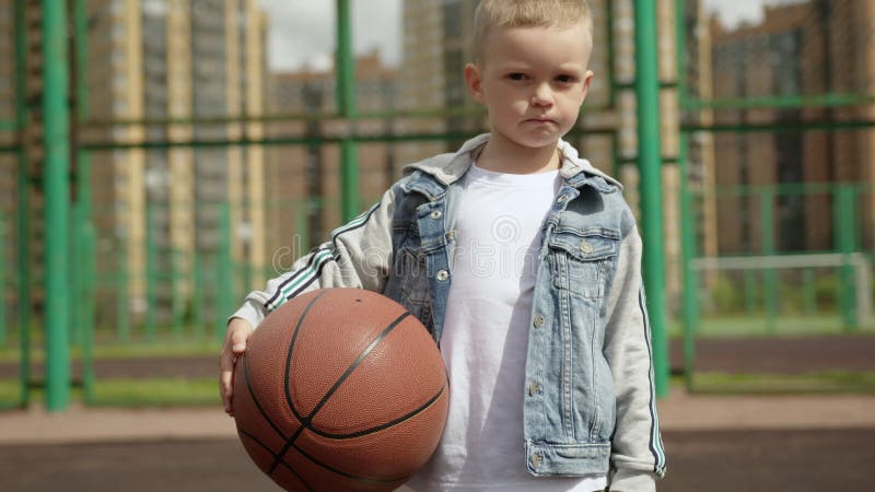 Retrato de niño de pie en una cancha de baloncesto sosteniendo una pelota en el distrito de la ciudad. almacen de metraje de vídeo