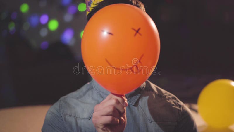Retrato de um homem novo seguro no chapéu do aniversário que remove um balão pintado com a cara engraçada que olha na câmera E