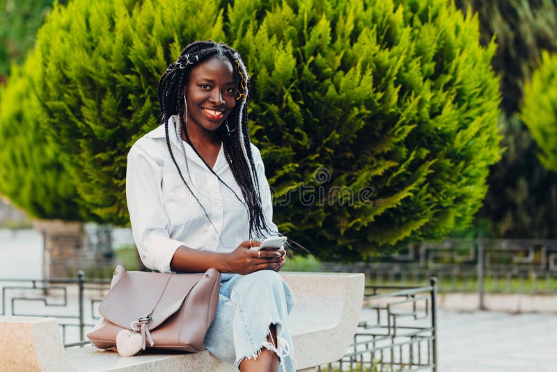 Retrato de uma jovem afro-americana sorridente com rabos-de-porco com um telefone sentado num banco fora num dia ensolarado Ao ar foto de stock Retrato de uma jovem afro-americana sorridente com rabos-de-porco com um telefone sentado num banco fora num dia ensolarado Ao ar foto de stock