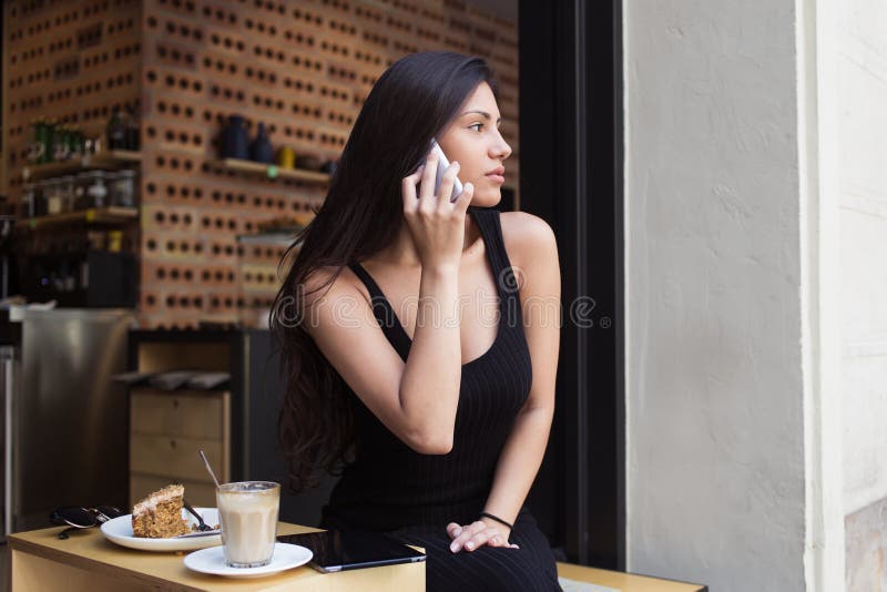 Retrato de uma jovem mulher bonita que fala no telefone celular ao relaxar na cafetaria durante o café da manhã imagens de stock Retrato de uma jovem mulher bonita que fala no telefone celular ao relaxar na cafetaria durante o café da manhã imagens de stock