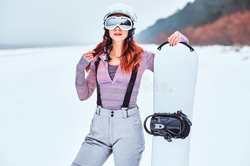 Retrato de uma menina bonita do ruivo no capacete protetor e dos óculos de proteção que guardam um snowboard, levantando em uma p imagens de stock royalty free Retrato de uma menina bonita do ruivo no capacete protetor e dos óculos de proteção que guardam um snowboard, levantando em uma p imagens de stock royalty free