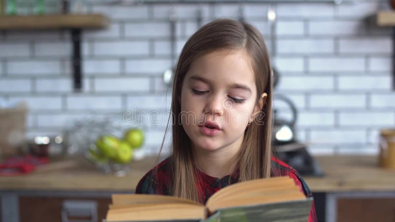 Retrato de uma menina bonito em uma camisa de manta que lê um livro que senta-se na mesa de cozinha