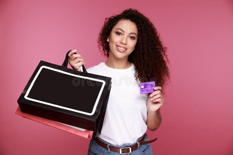 Retrato de uma mulher africana nova entusiasmado que guarda sacos de compras e que mostra o cartão de crédito isolado sobre o fun fotos de stock Retrato de uma mulher africana nova entusiasmado que guarda sacos de compras e que mostra o cartão de crédito isolado sobre o fun fotos de stock