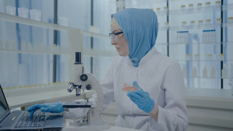 Retrato de uma mulher cientista muçulmana, descontente e intrigada, em um casaco hijab e branco olhando através de um microscópio