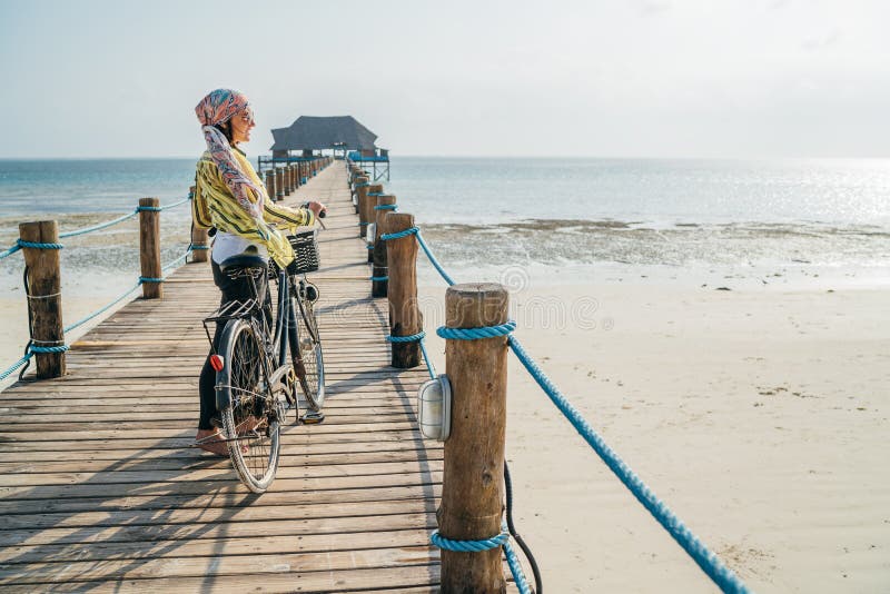 Portrait of a smiling woman dressed in light summer clothes, sunglasses with bicycle on the wooden sea pier on the sandy Zanzibar beach.Careless vacation in the tropical countries concept image. Portrait of a smiling woman dressed in light summer clothes, sunglasses with bicycle on the wooden sea pier on the sandy Zanzibar beach.Careless vacation in the tropical countries concept image
