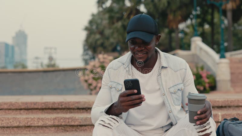 Retrato de un afroamericano sonriente con café usando un smartphone sentado en las escaleras del parque almacen de metraje de vídeo