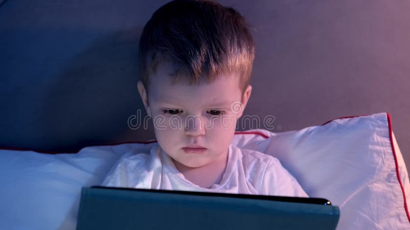 Retrato de un chico caucásico rubio lindo mirando caricatura en una tableta sentada en la cama a la luz de la luna desde la ventan almacen de video