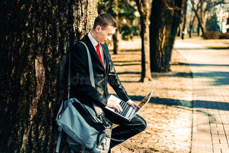 Retrato de un estudiante en un traje negro y del lazo rojo en la naturaleza cerca de un árbol grande en el hombro de un bolso jov foto de archivo libre de regalías Retrato de un estudiante en un traje negro y del lazo rojo en la naturaleza cerca de un árbol grande en el hombro de un bolso jov foto de archivo libre de regalías