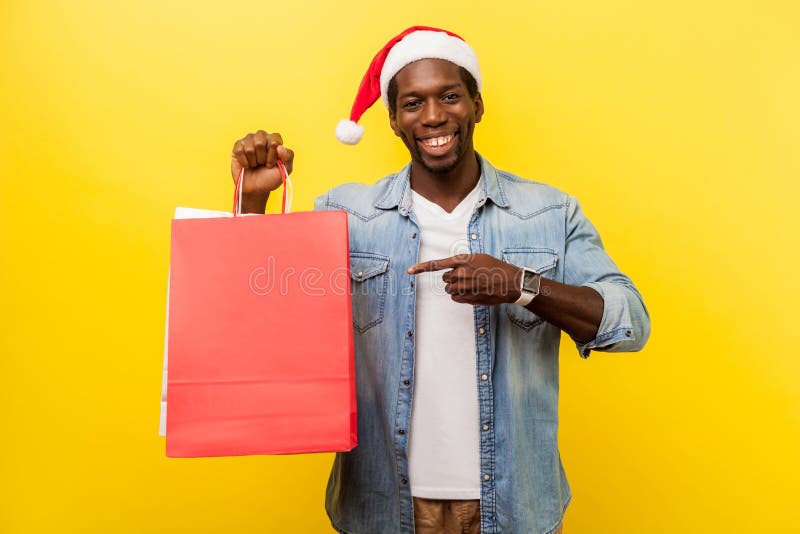 Retrato de un excitado y apuesto joven con sombrero de santa y camisa informal de denim que apunta a la bolsa de compras y mira l imágenes de archivo libres de regalías Retrato de un excitado y apuesto joven con sombrero de santa y camisa informal de denim que apunta a la bolsa de compras y mira l imágenes de archivo libres de regalías
