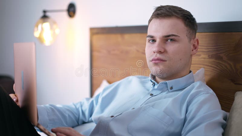Retrato de un joven inteligente con una laptop en la cama y mirando la cámara con expresión facial segura