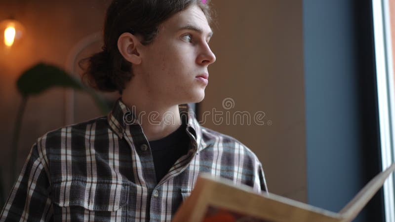 Retrato de un joven inteligente leyendo un libro en cámara lenta mirando por la ventana. caucásico de confianza sentado