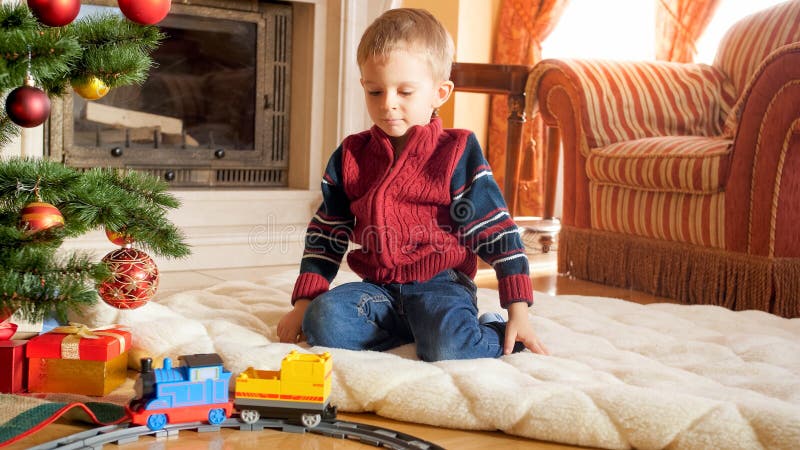 Retrato de un niño pequeño sentado en el suelo jugando con un tren de juguete que Santa le dio por Navidad. Niños que reciben imágenes de archivo libres de regalías Retrato de un niño pequeño sentado en el suelo jugando con un tren de juguete que Santa le dio por Navidad. Niños que reciben imágenes de archivo libres de regalías