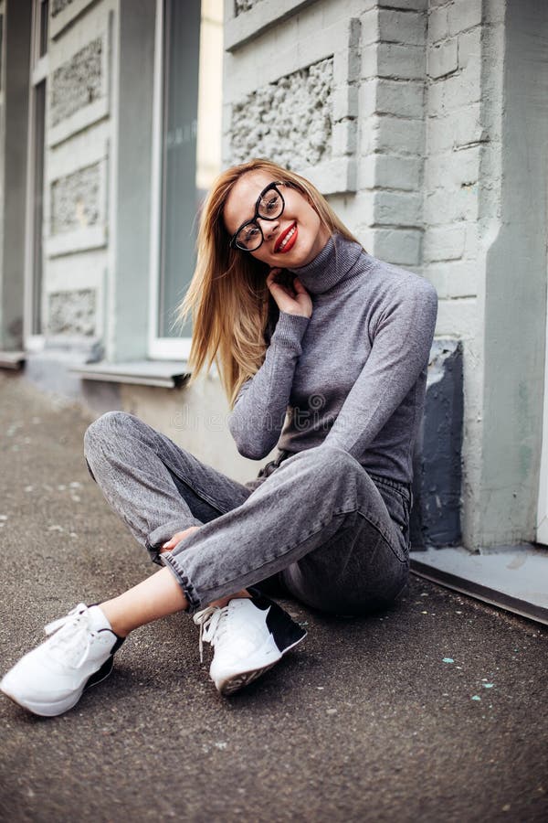 Retrato De Una Alegre Mujer Blanca En Gafas, Usando Jeans Grises Y Suéter Gris Sentado En El Pavimento Y Riendo Foto de archivo - Imagen de ciudad, 160543556