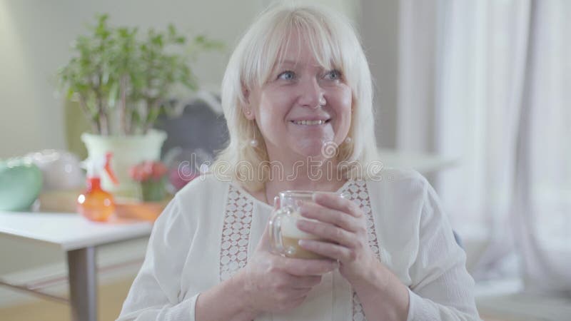 Retrato de una atractiva mujer caucásica con pelo rubio y ojos grises bebiendo café y sonriendo Feliz jubilado metrajes