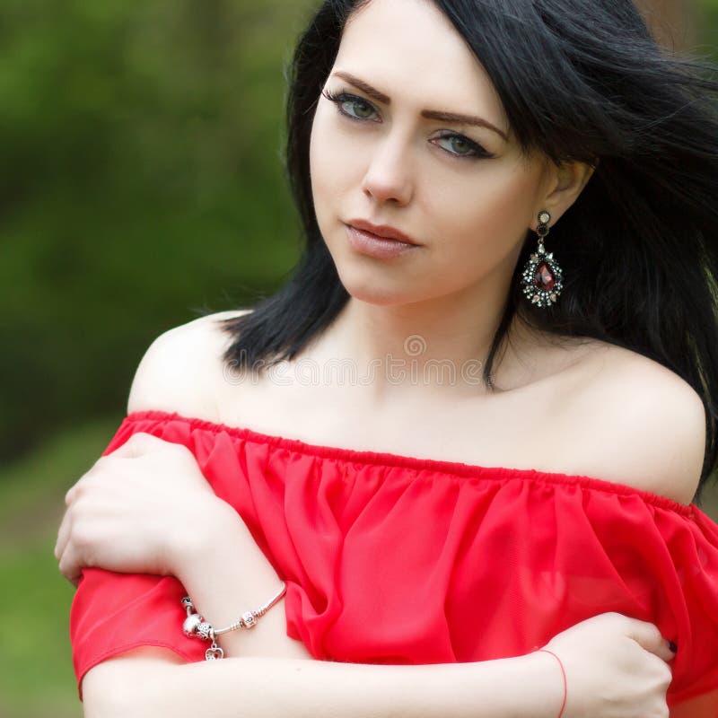 Retrato de una chica joven hermosa con los ojos verdes y los hombros desnudos, en un vestido rojo en una naturaleza verde del ver foto de archivo libre de regalías Retrato de una chica joven hermosa con los ojos verdes y los hombros desnudos, en un vestido rojo en una naturaleza verde del ver foto de archivo libre de regalías