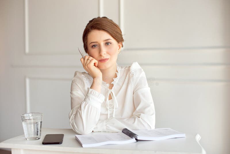Retrato de una empresaria morena joven sonriente hermosa en una camisa blanca que se sienta en un puesto de trabajo moderno brill imagenes de archivo Retrato de una empresaria morena joven sonriente hermosa en una camisa blanca que se sienta en un puesto de trabajo moderno brill imagenes de archivo