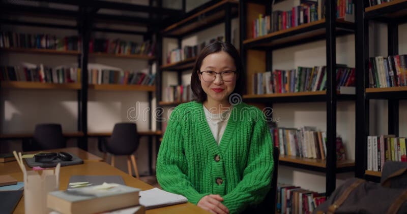 Retrato de una feliz chica asiática con gafas y un suéter verde sentada en una mesa en la biblioteca almacen de metraje de vídeo