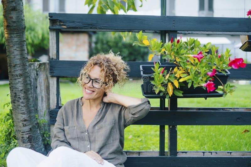 Portrait of young mature woman having relax and enjoying home outdoor garden sitting and smiling. One female people serene relaxing outside home. Happy adult lady wearing eyeglasses sit down. Cheerful. Portrait of young mature woman having relax and enjoying home outdoor garden sitting and smiling. One female people serene relaxing outside home. Happy adult lady wearing eyeglasses sit down. Cheerful
