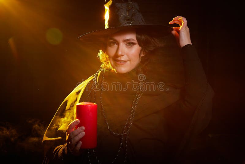 Retrato de una linda bruja joven con un sombrero puntiagudo con la varita de la vela en sus manos y fondo negro. niña o mujer en imagen de archivo