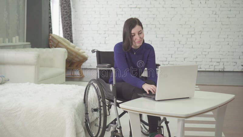 Retrato de una mujer discapacitada hermosa joven sonriente en una silla de ruedas, trabajando en casa en un ordenador portátil, t