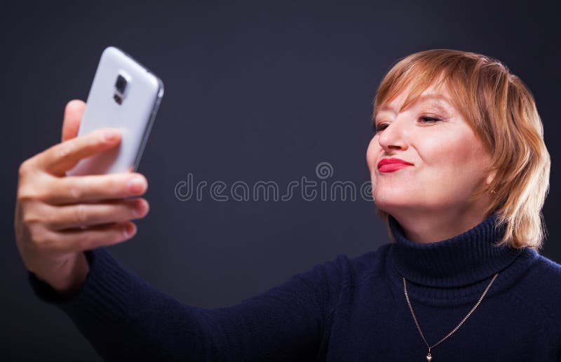 Retrato de una mujer de mediana edad que hace la foto del selfie con smartphone en un fondo negro imagenes de archivo Retrato de una mujer de mediana edad que hace la foto del selfie con smartphone en un fondo negro imagenes de archivo