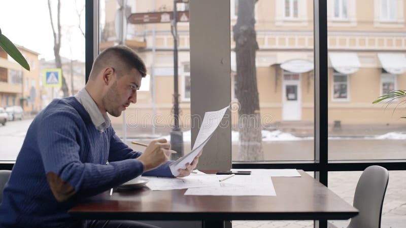 Retrato del arquitecto de sexo masculino joven que trabaja en café cerca de la ventana y de los papeles que lanzan lejos