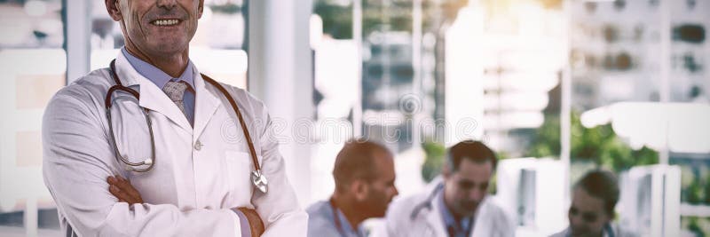 Retrato del doctor feliz que se coloca con los brazos cruzados mientras que sus colegas trabajan fotografía de archivo libre de regalías Retrato del doctor feliz que se coloca con los brazos cruzados mientras que sus colegas trabajan fotografía de archivo libre de regalías