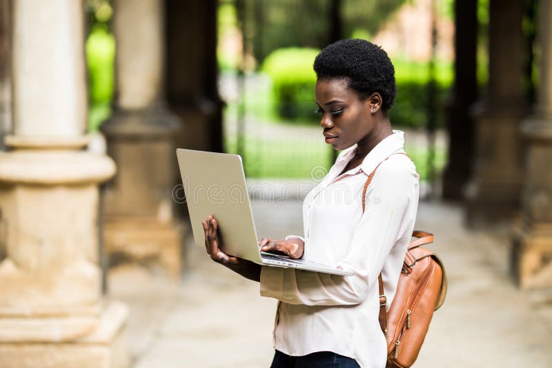 Retrato del estudiante afroamericano sonriente que hace la preparación al aire libre usando el ordenador portátil para la investi foto de archivo Retrato del estudiante afroamericano sonriente que hace la preparación al aire libre usando el ordenador portátil para la investi foto de archivo