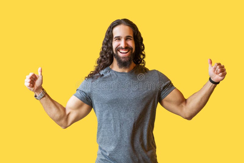 Retrato del hombre joven barbudo feliz con el pelo rizado largo en la situación gris de la camiseta con los pulgares para arriba