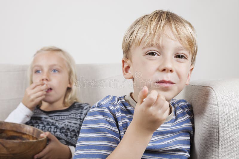 Retrato del muchacho joven con la hermana que ve la TV y que come las palomitas imagen de archivo Retrato del muchacho joven con la hermana que ve la TV y que come las palomitas imagen de archivo