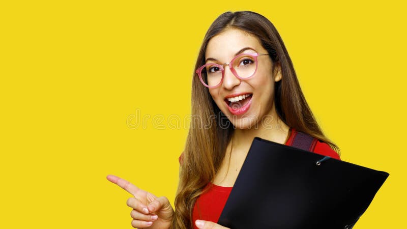 Retrato do estudante fêmea do cabelo longo bonito que veste vidros vermelhos do t-shirt e do lerdo, apontando com o indicador no foto de stock Retrato do estudante fêmea do cabelo longo bonito que veste vidros vermelhos do t-shirt e do lerdo, apontando com o indicador no foto de stock