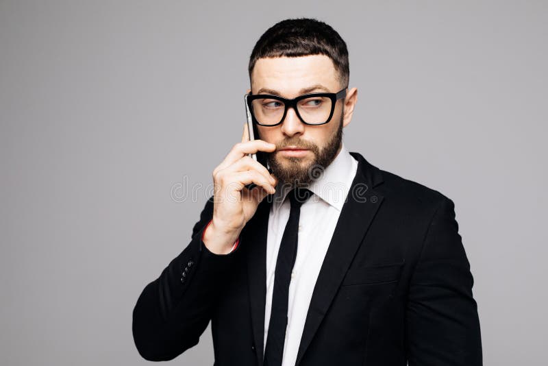 Retrato do homem atrativo novo sério no terno preto que fala no telefone celular no fundo cinzento imagens de stock Retrato do homem atrativo novo sério no terno preto que fala no telefone celular no fundo cinzento imagens de stock
