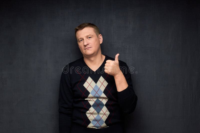 Studio half-length portrait of caucasian man wearing sweater, raising thumb up in approval and like gesture, but with indifferent expression, looking with displeasure at camera, over gray background. Studio half-length portrait of caucasian man wearing sweater, raising thumb up in approval and like gesture, but with indifferent expression, looking with displeasure at camera, over gray background