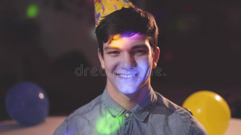 Retrato do homem novo de sorriso no chapéu do aniversário que olha na câmera Luzes brilhantes de cores diferentes em sua cara e
