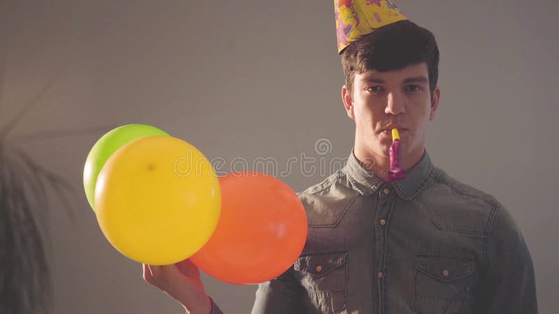 Retrato do homem novo no chapéu do aniversário com o noisemaker na boca que olha na câmera que guarda balões Luzes brilhantes de